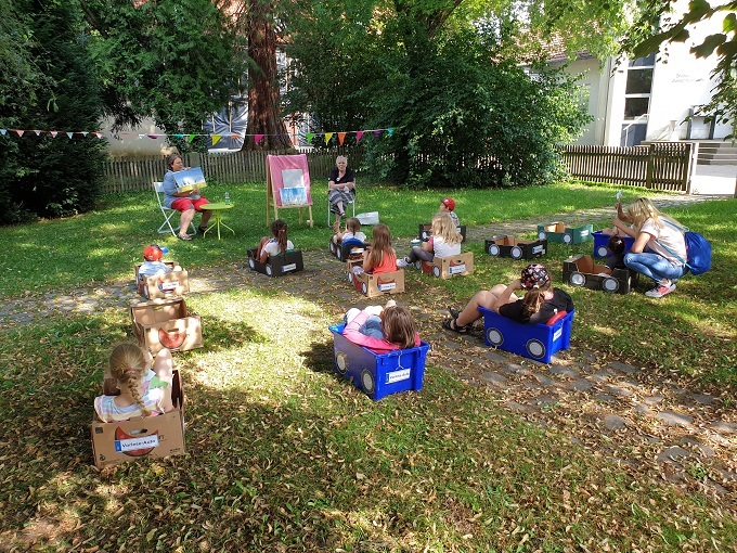 Kinder sitzen in bemalten Kartons auf einer Wiese und bekommen vorgelesen
