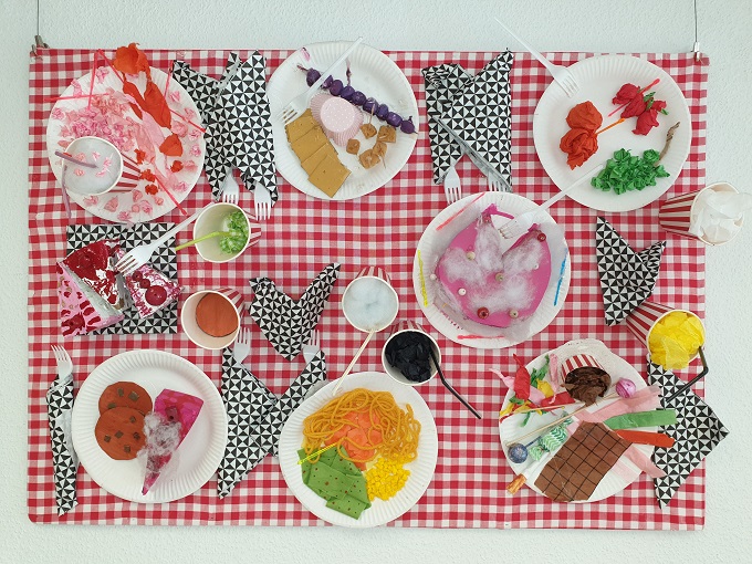 Ein Wandteppich als rotkarierte Picknickdecke mit Tellern mit Essen, Bechern mit Trinken und Besteck in Servietten gestaltet